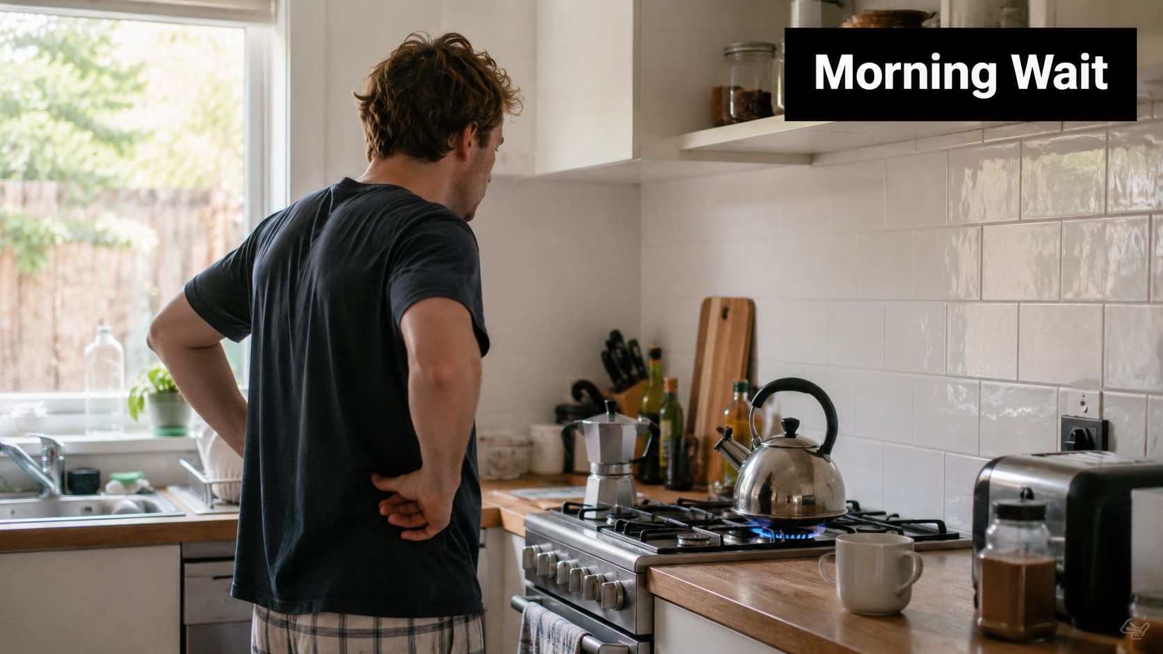 A man in pajamas stands in his kitchen waiting for the kettle to boil on the stove.
